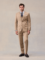Model facing forward in a camel three-piece suit with notch lapels, navy tie and white pocket square.
