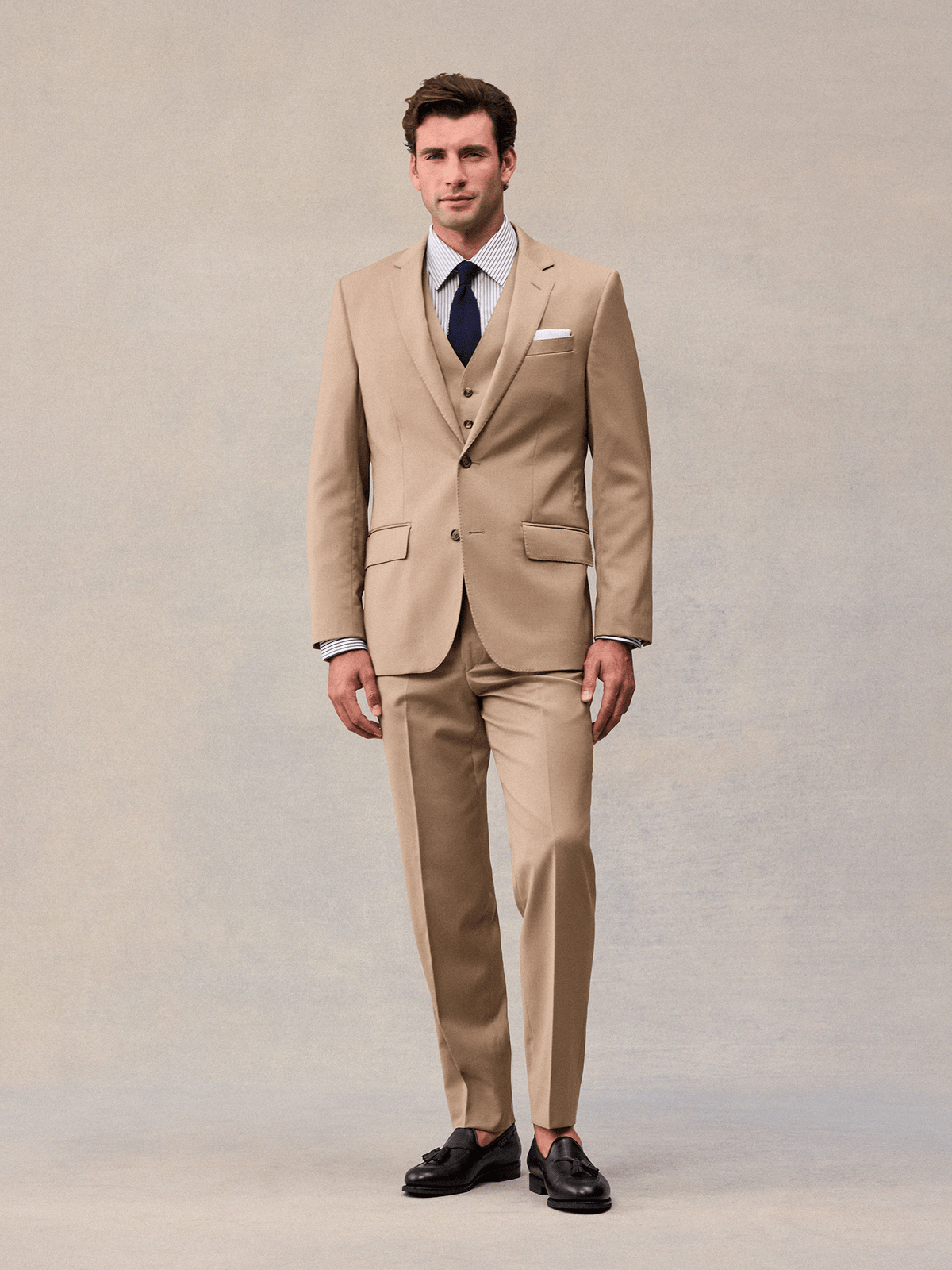 Model facing forward in a camel three-piece suit with notch lapels, navy tie and white pocket square.