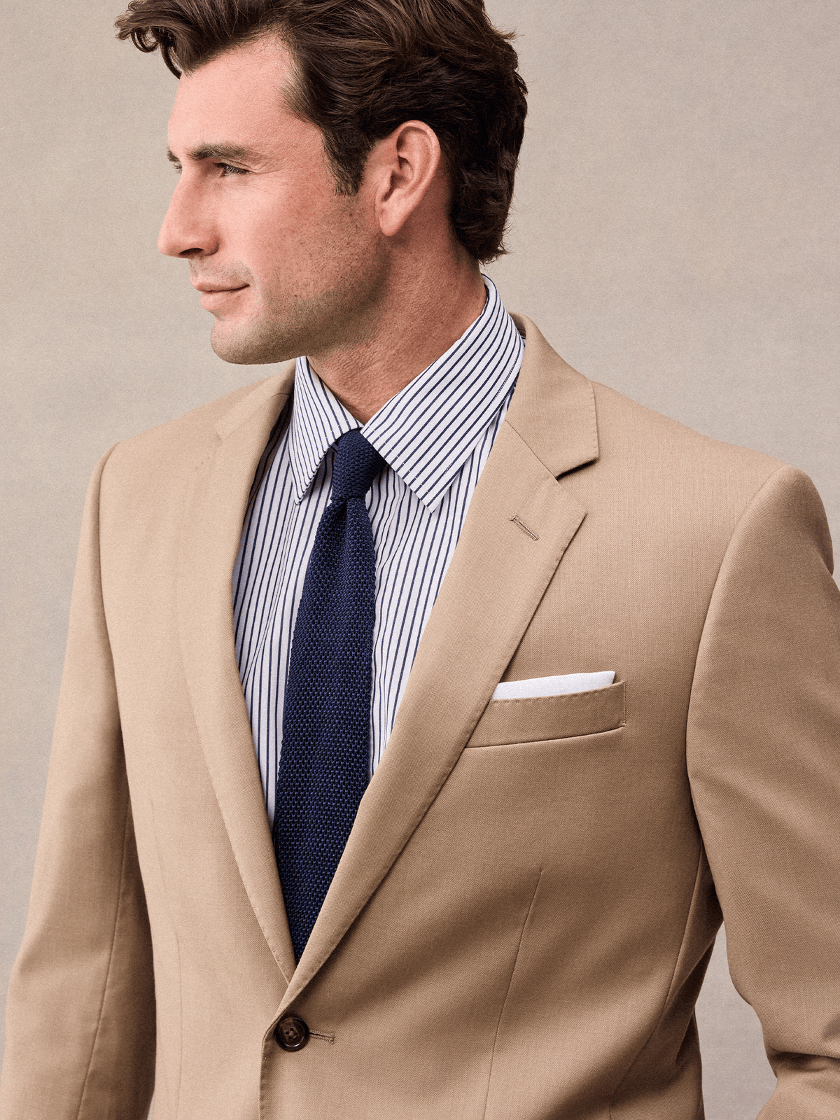 Close-up of a camel suit jacket's notch lapel with white pocket square and navy knit tie.