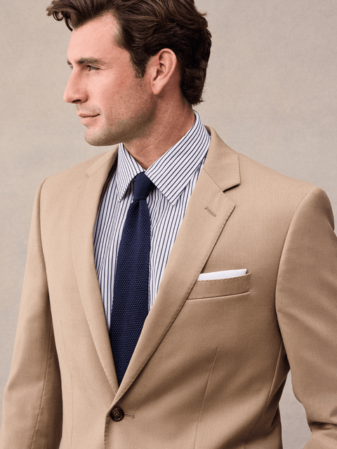 Close-up of a camel suit jacket's notch lapel with white pocket square and navy knit tie.