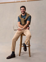 Man in slate blue pique polo and tan chinos, seated on stool with sweater tied around shoulders