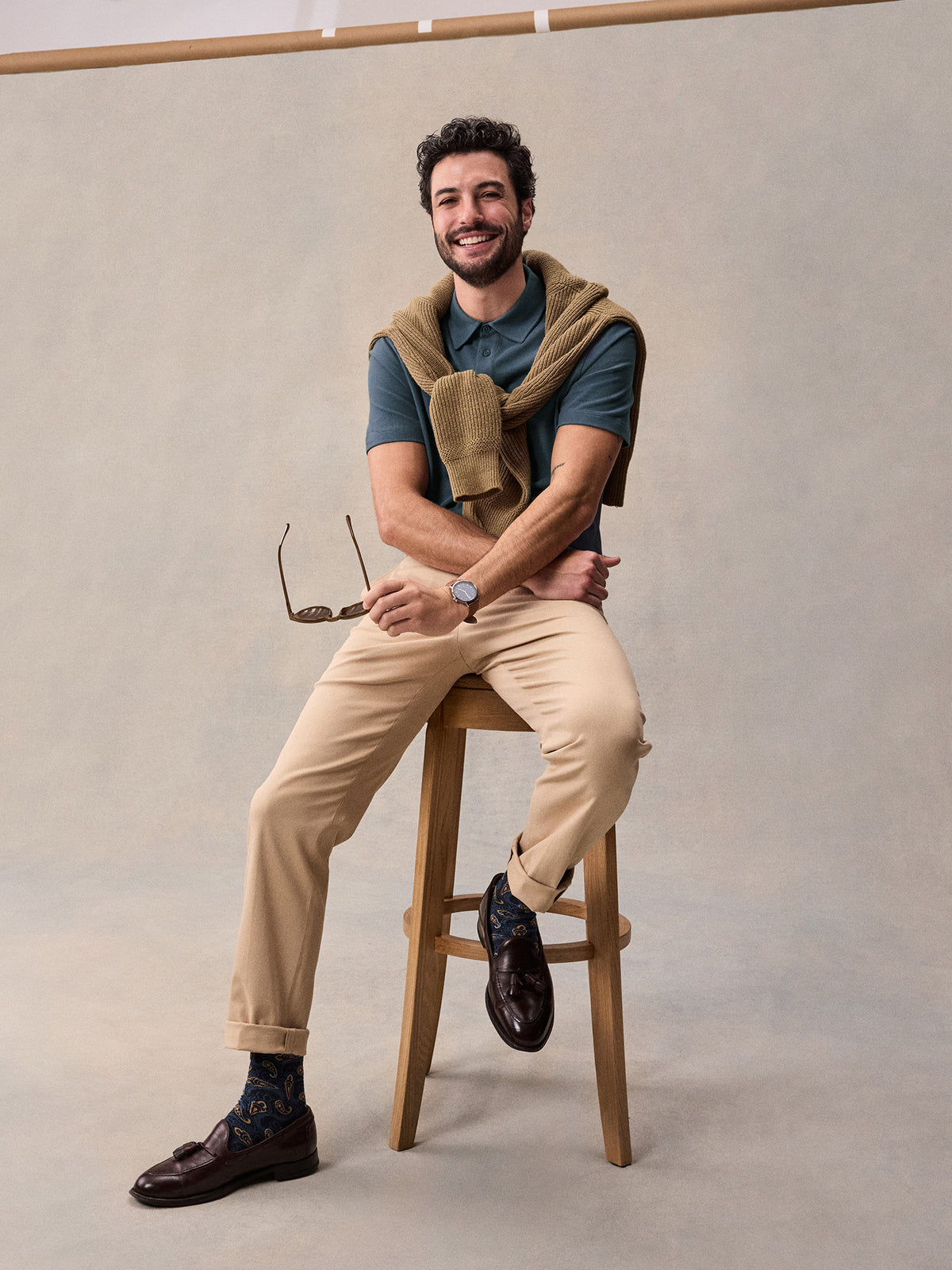 Man in slate blue pique polo and tan chinos, seated on stool with sweater tied around shoulders