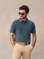 Man in slate blue pique polo and tan chinos wearing sunglasses, hands in pockets