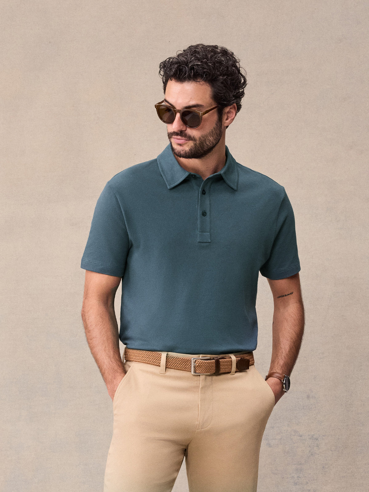 Man in slate blue pique polo and tan chinos wearing sunglasses, hands in pockets