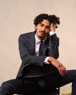 Man in a dark suit sitting on a chair with one hand on his head.