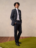 Man in a dark grey suit with a white shirt and black tie, standing on a green rug.