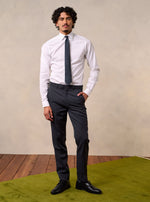 Man wearing a white shirt, dark tie, and grey pants, standing on a green rug.