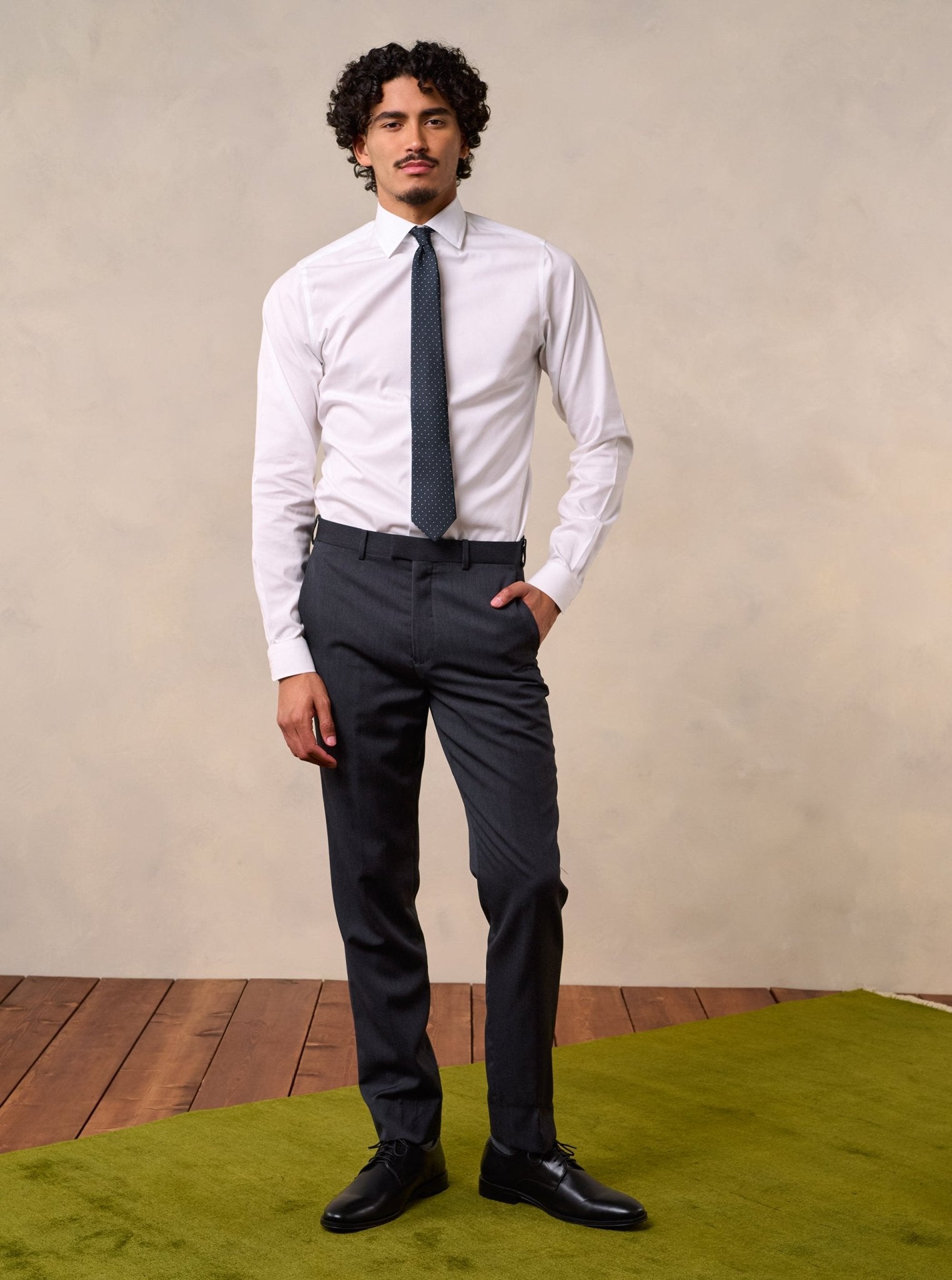 Man wearing a white shirt, dark tie, and grey pants, standing on a green rug.