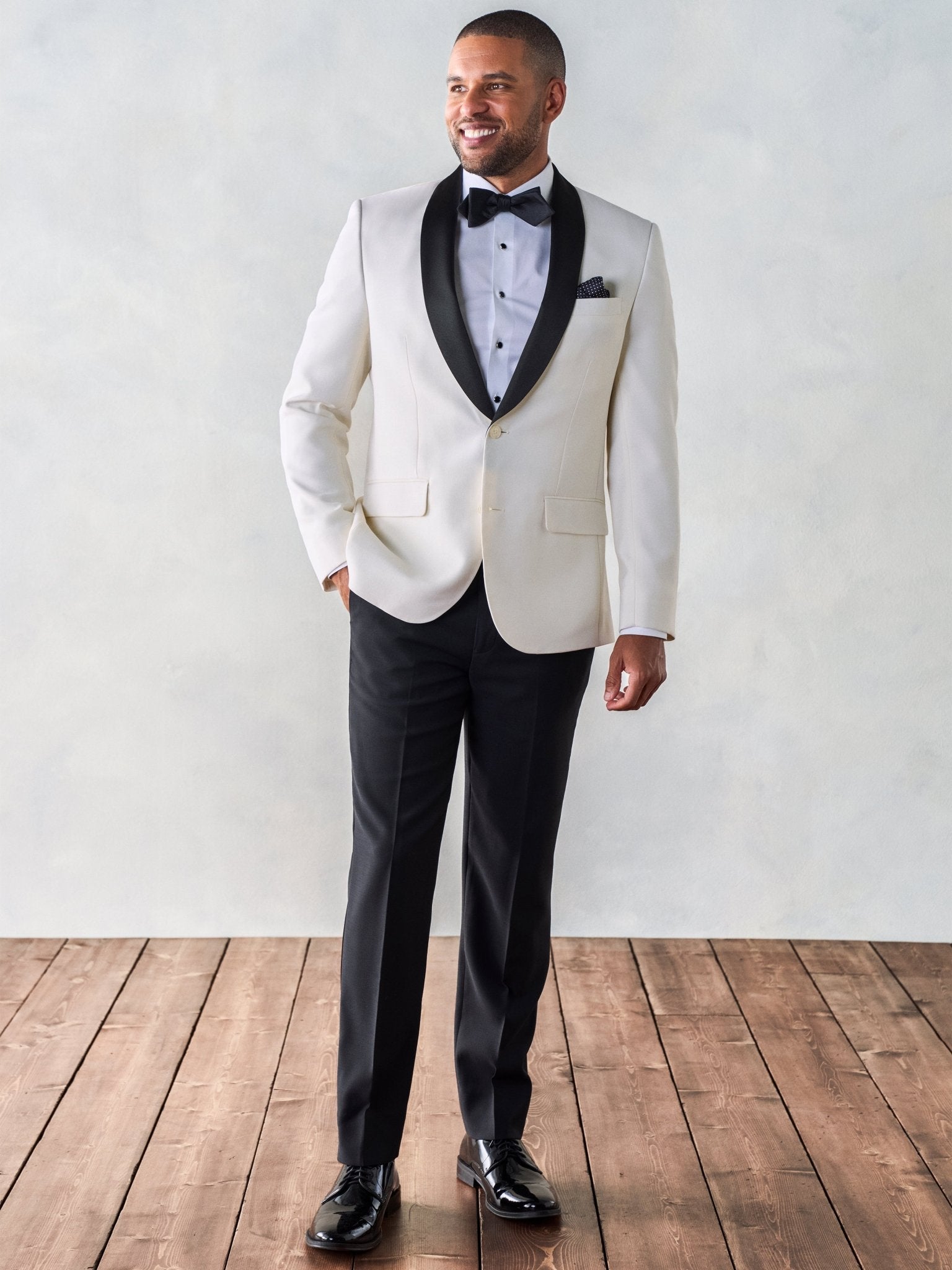 Man in a white tuxedo jacket, black bow tie, and black pants, standing on wooden floor.