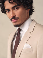 Man in a beige suit with a brown tie and white pocket square, looking directly at the camera.