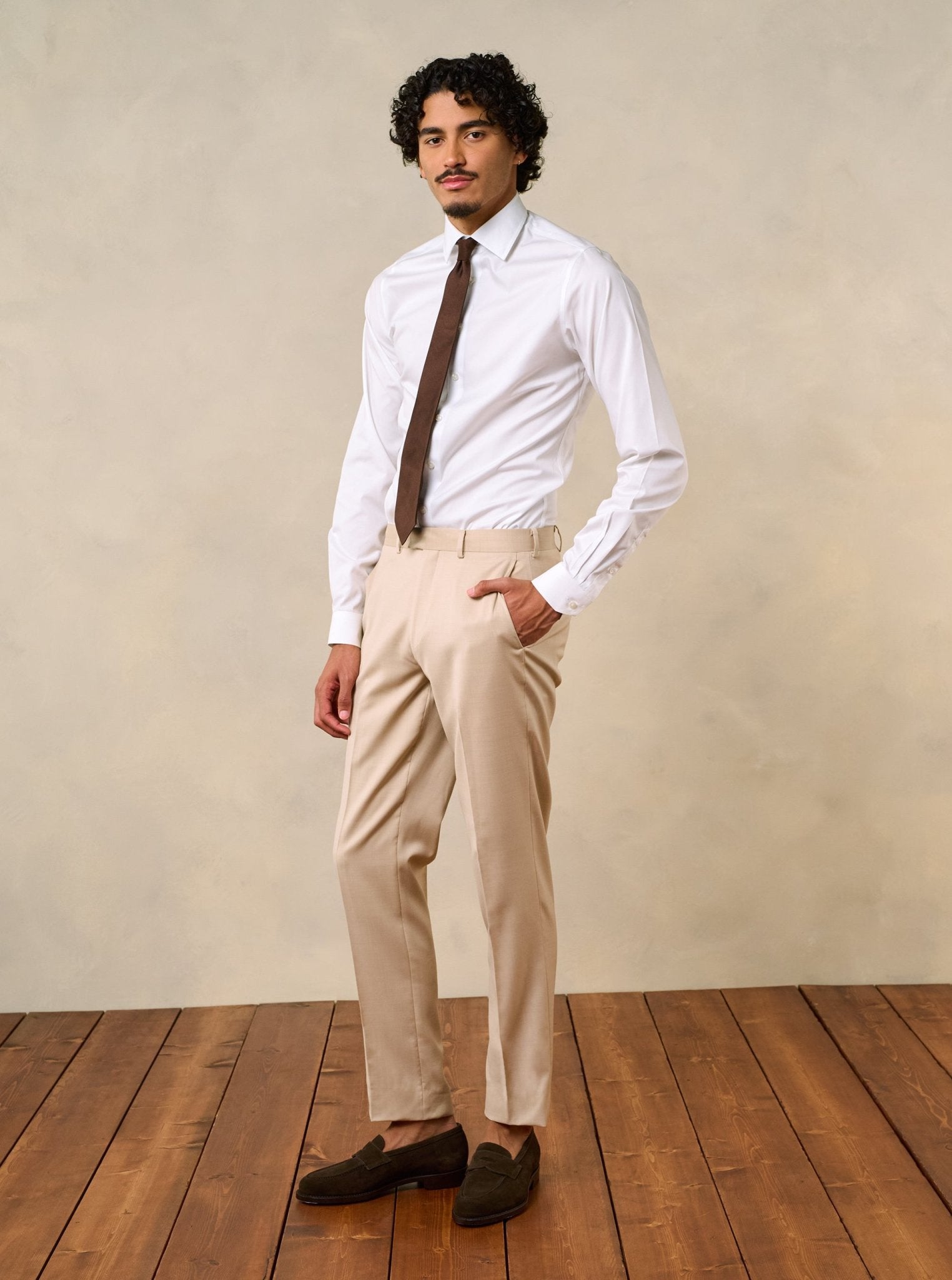 Man wearing a white shirt, brown tie, and beige pants, standing on wooden floor.
