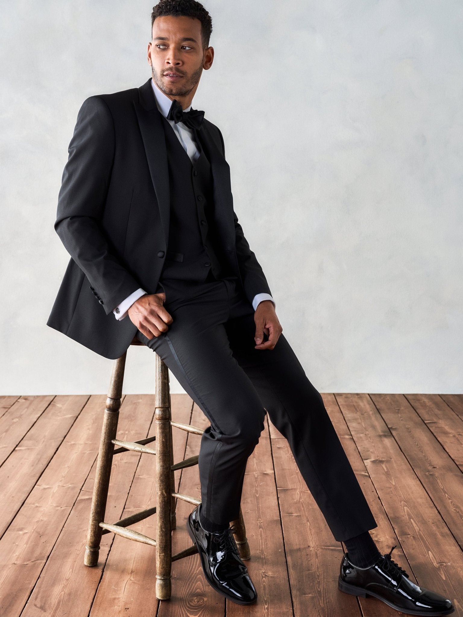 Man in a black tuxedo sitting on a wooden stool.
