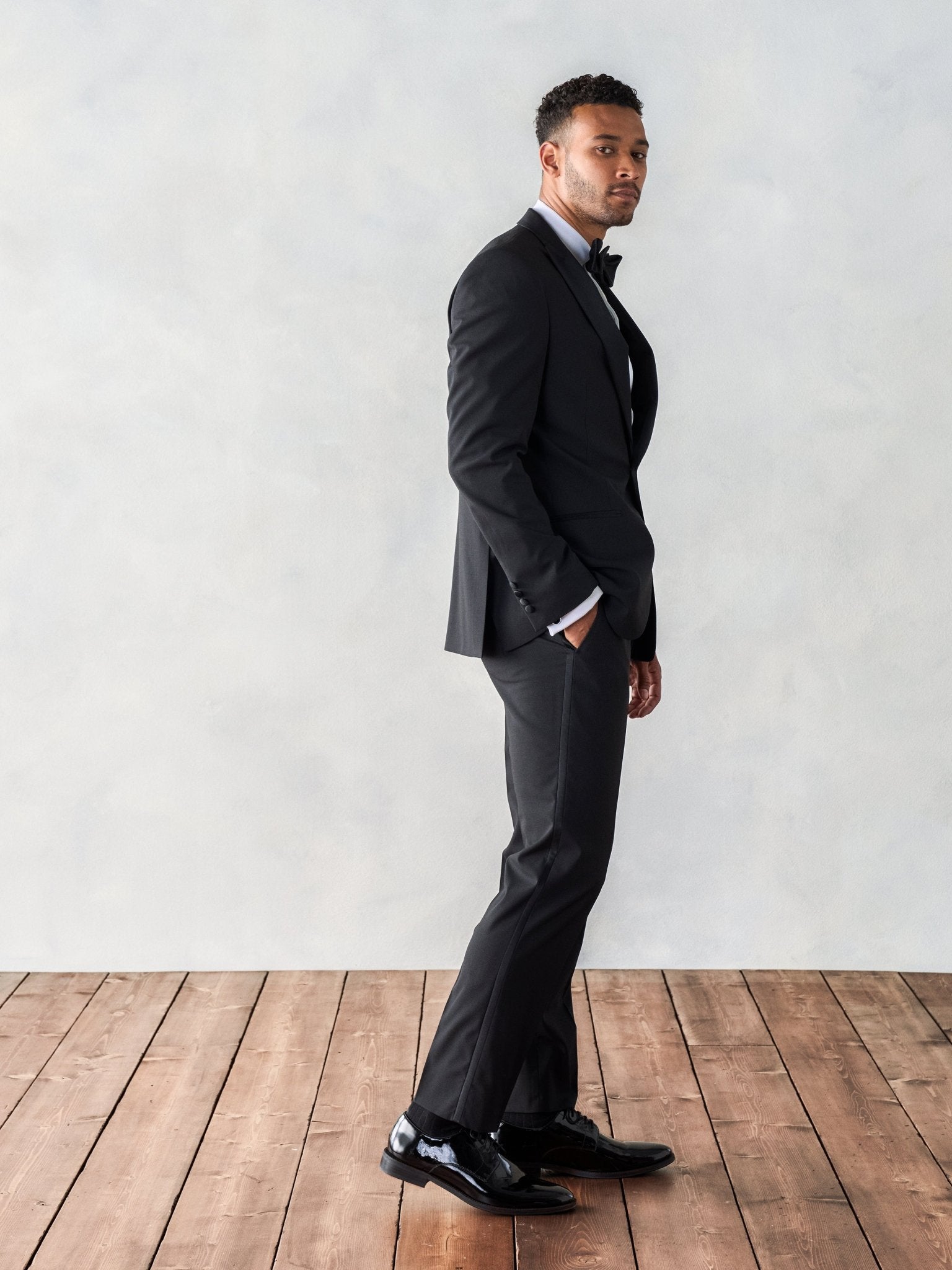 Man in a black tuxedo standing on wooden floor against a light background.