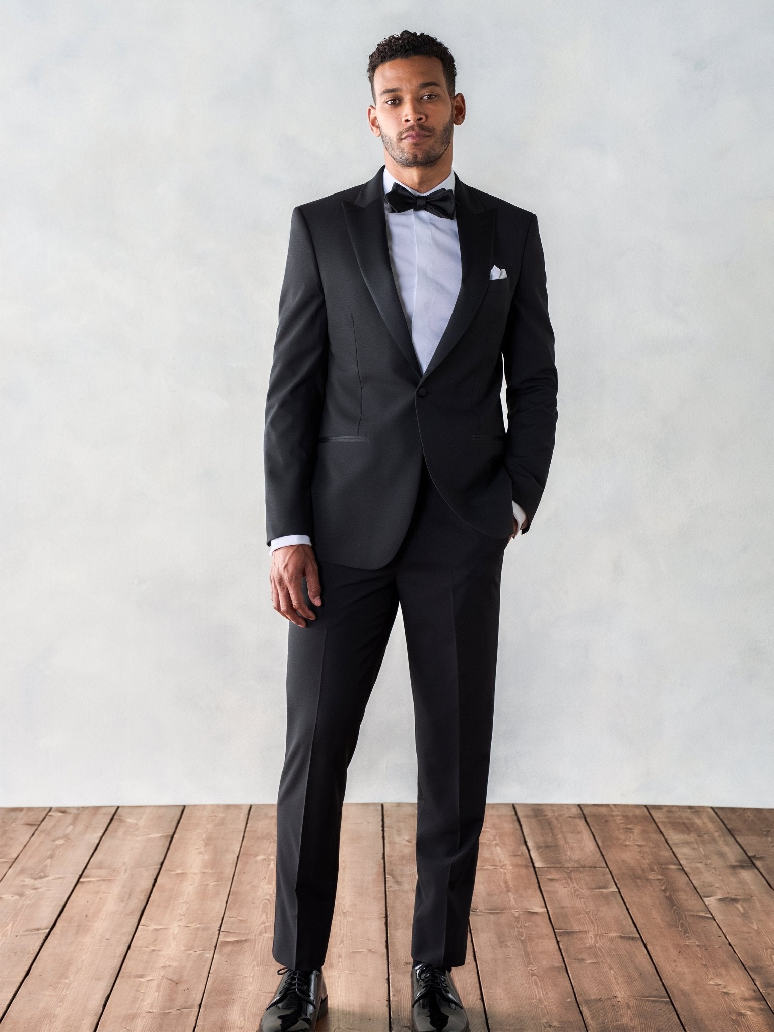 Man wearing a black tuxedo with a bow tie, standing on wooden floor.