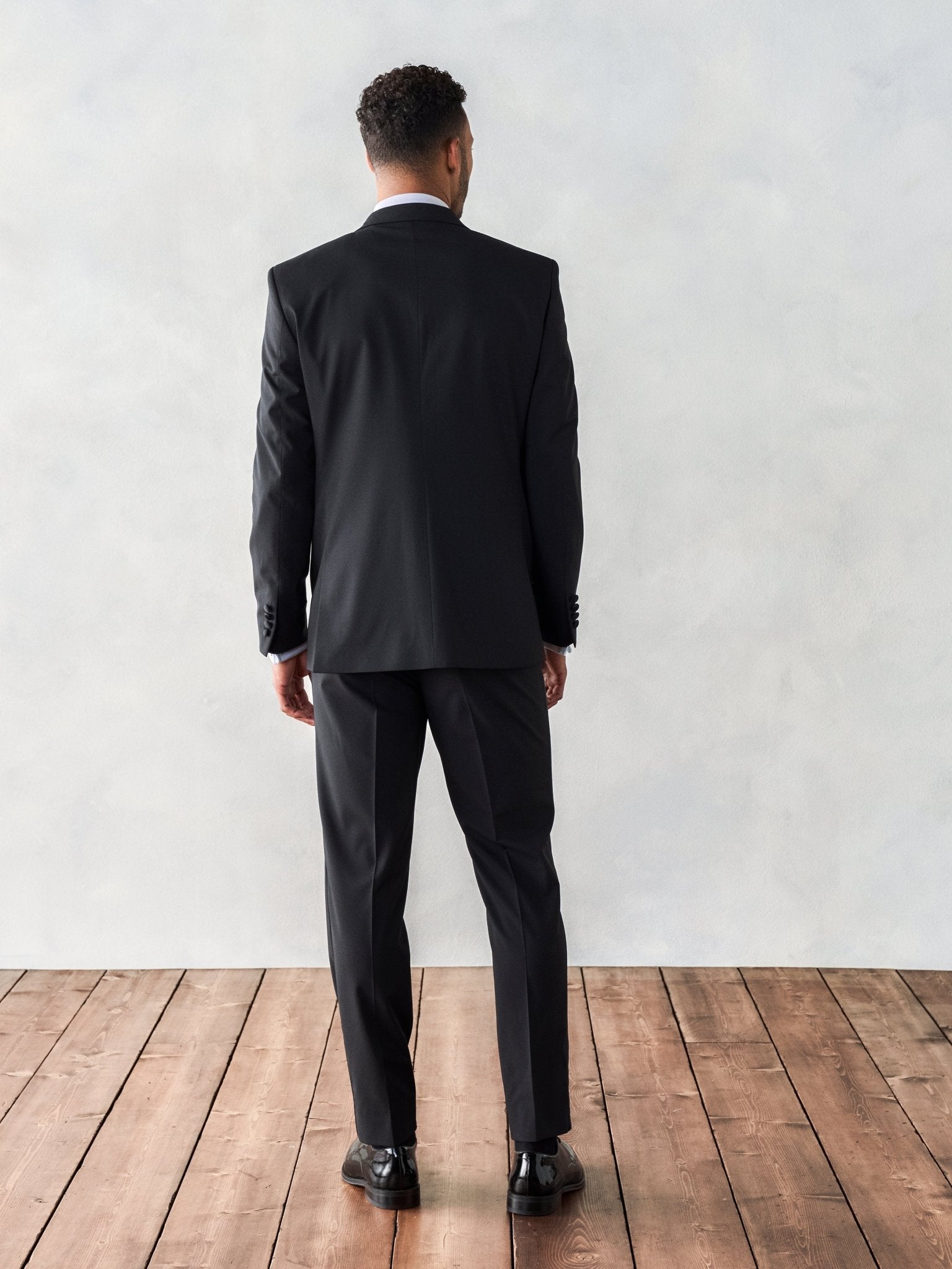 Man in a black tuxedo viewed from the back, standing on wooden floor.
