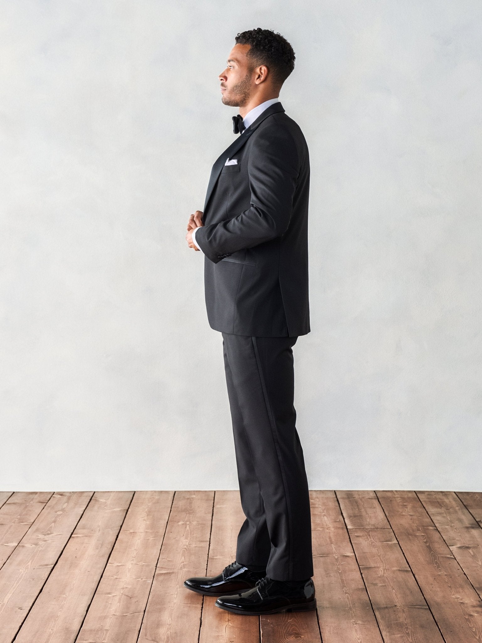 Man in a black tuxedo standing sideways on wooden floor.