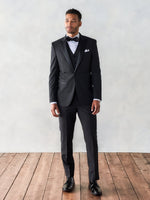 Man wearing a black tuxedo with a bow tie and dress shoes, standing on wooden floor.