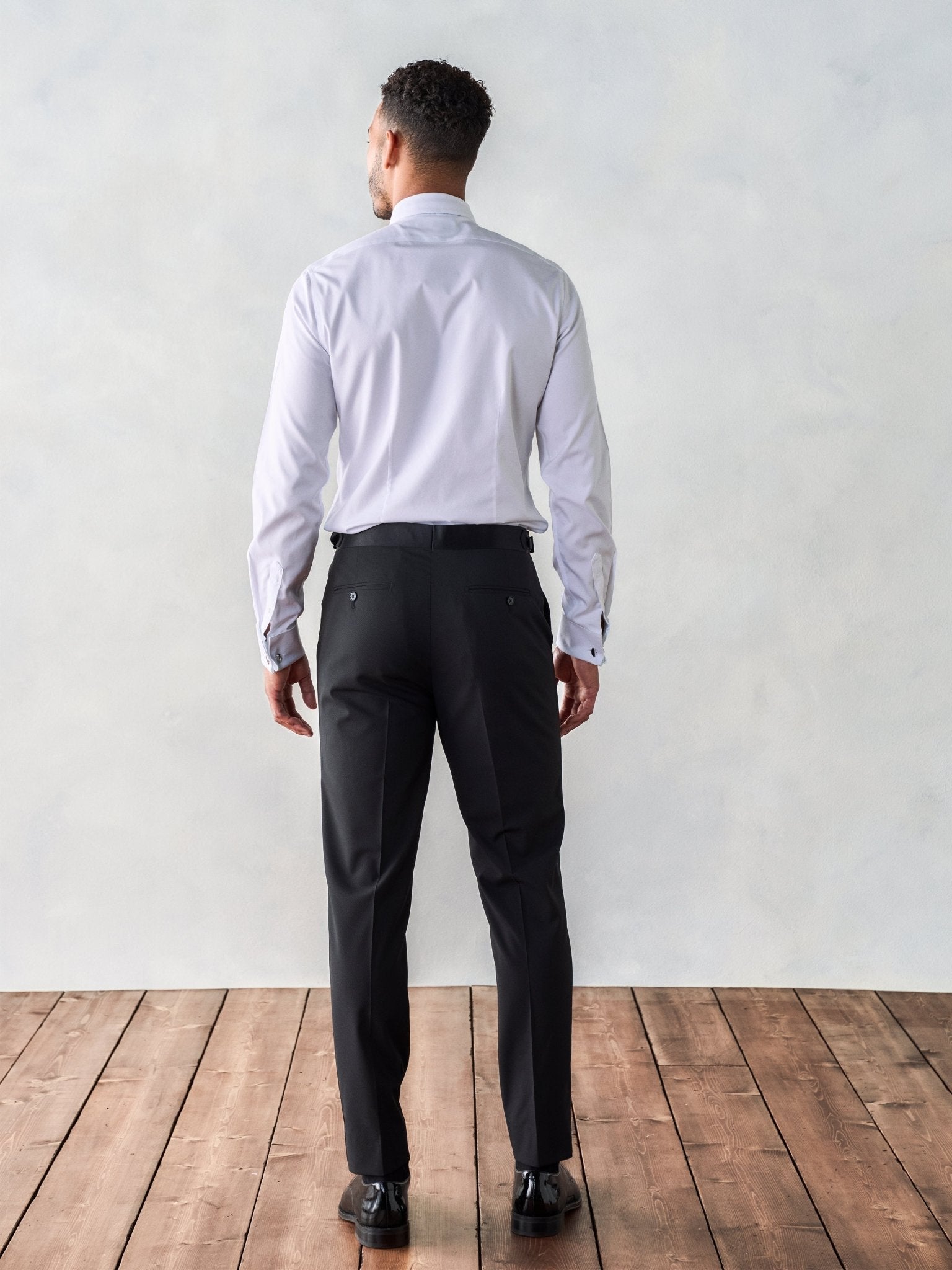 Man in a white shirt and black pants, viewed from behind, standing on wooden floor.