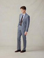 A man in a grey suit with a blue shirt and brown tie, standing against a neutral background.