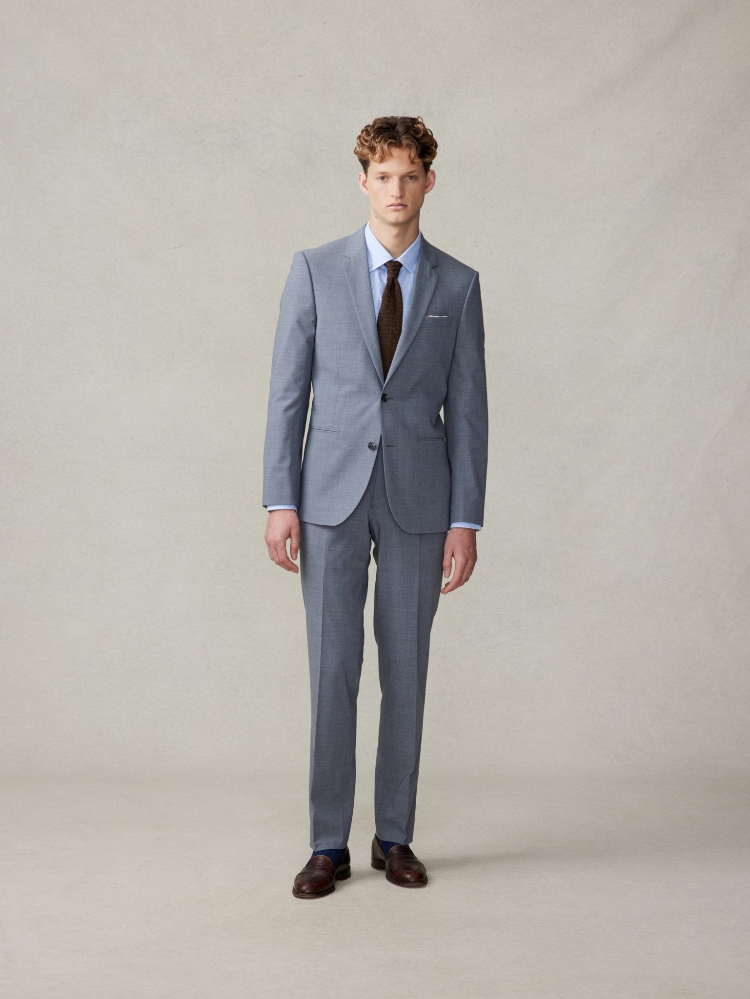 Man wearing a grey suit with a brown tie and brown shoes, standing against a neutral background.