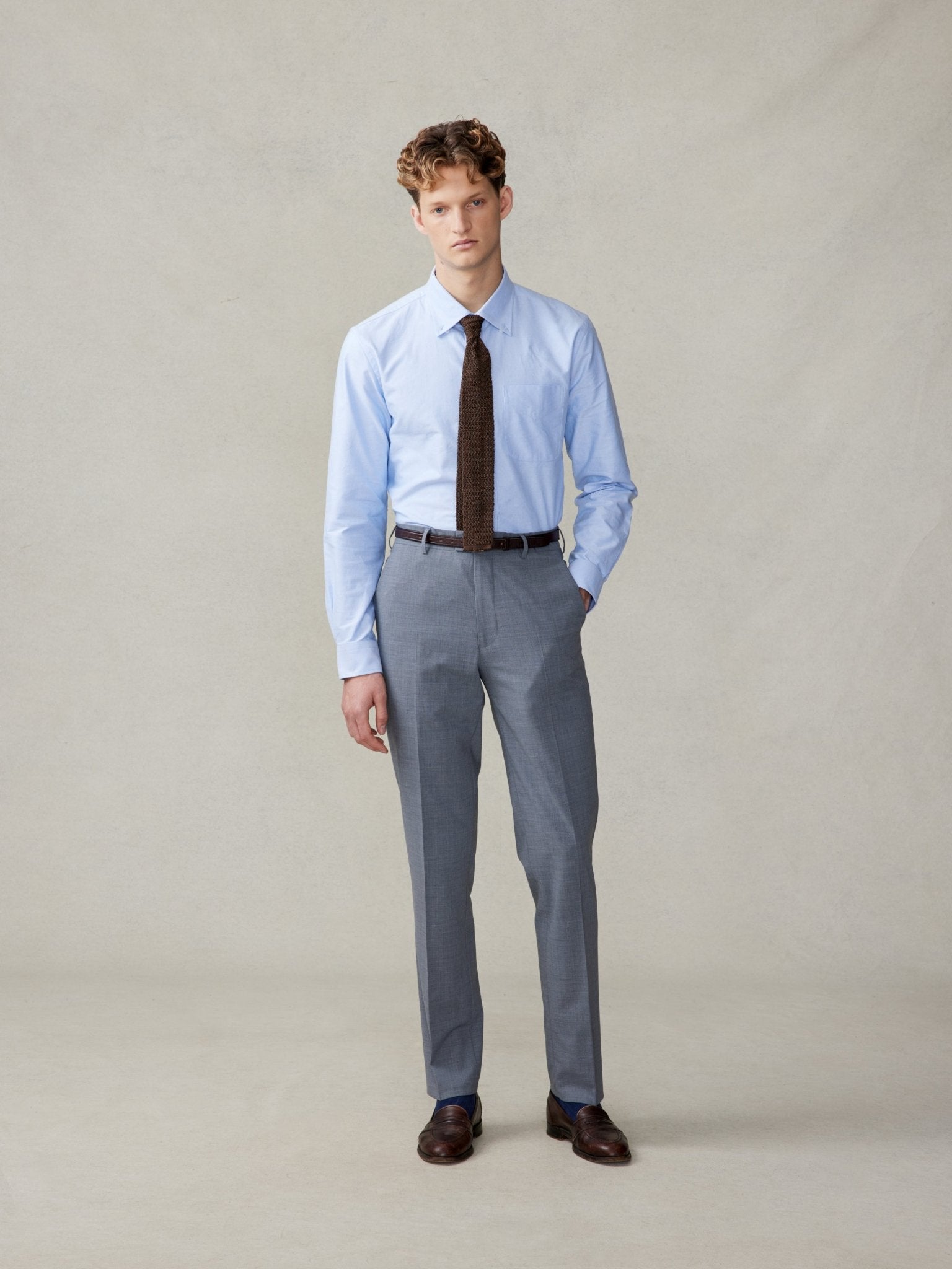 Man wearing a light blue shirt, brown tie, and gray trousers, standing against a neutral background.