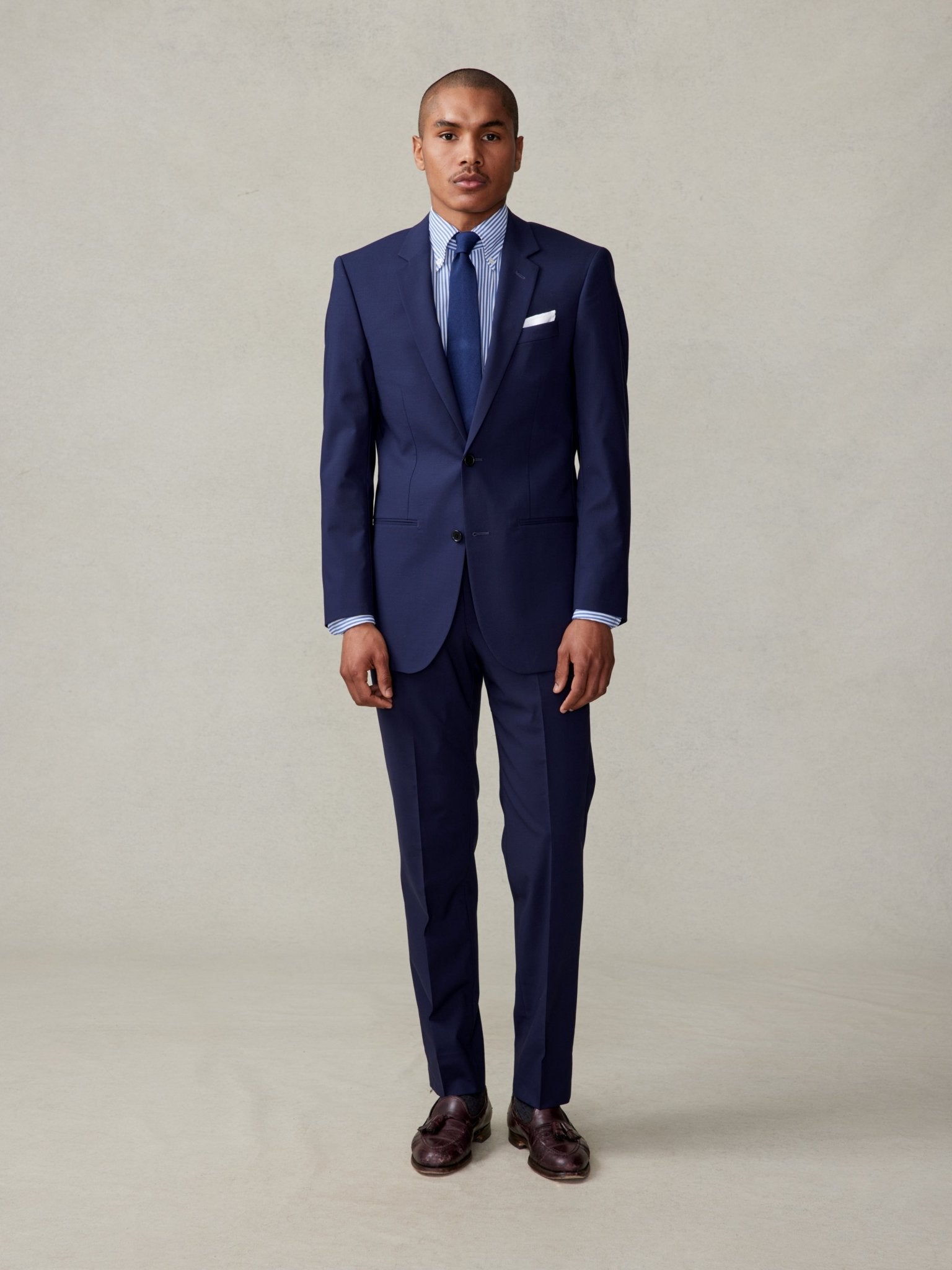 Man wearing a navy suit with a striped shirt and tie, standing on a neutral background.