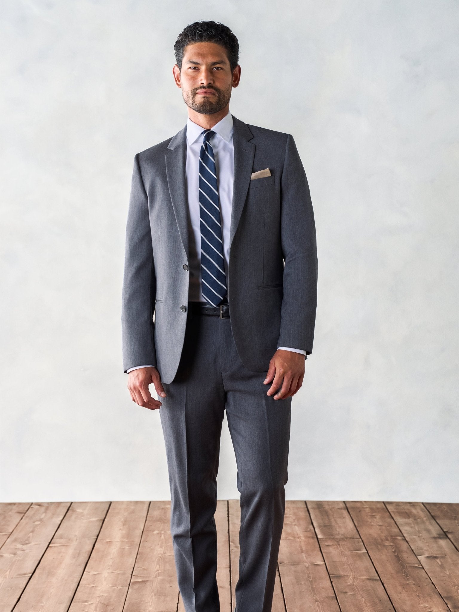 Man in a grey suit with a striped tie standing on wooden floor.