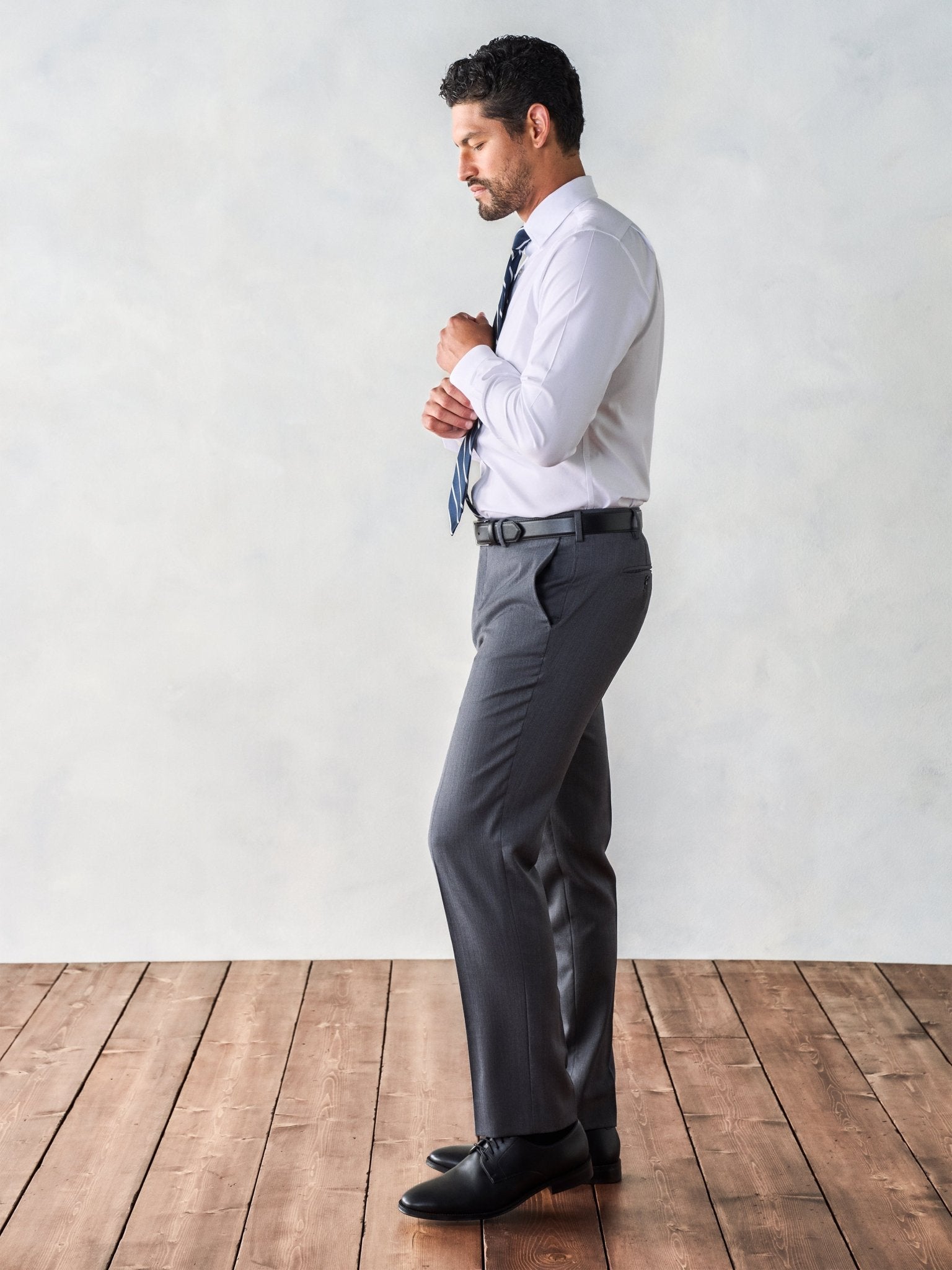 Man adjusting his shirt cuffs while wearing a white shirt and grey trousers.
