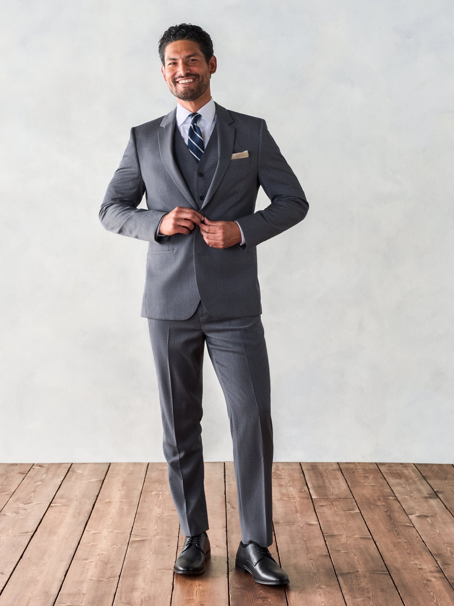 Man in a grey suit adjusting his jacket, smiling, standing on wooden floor.