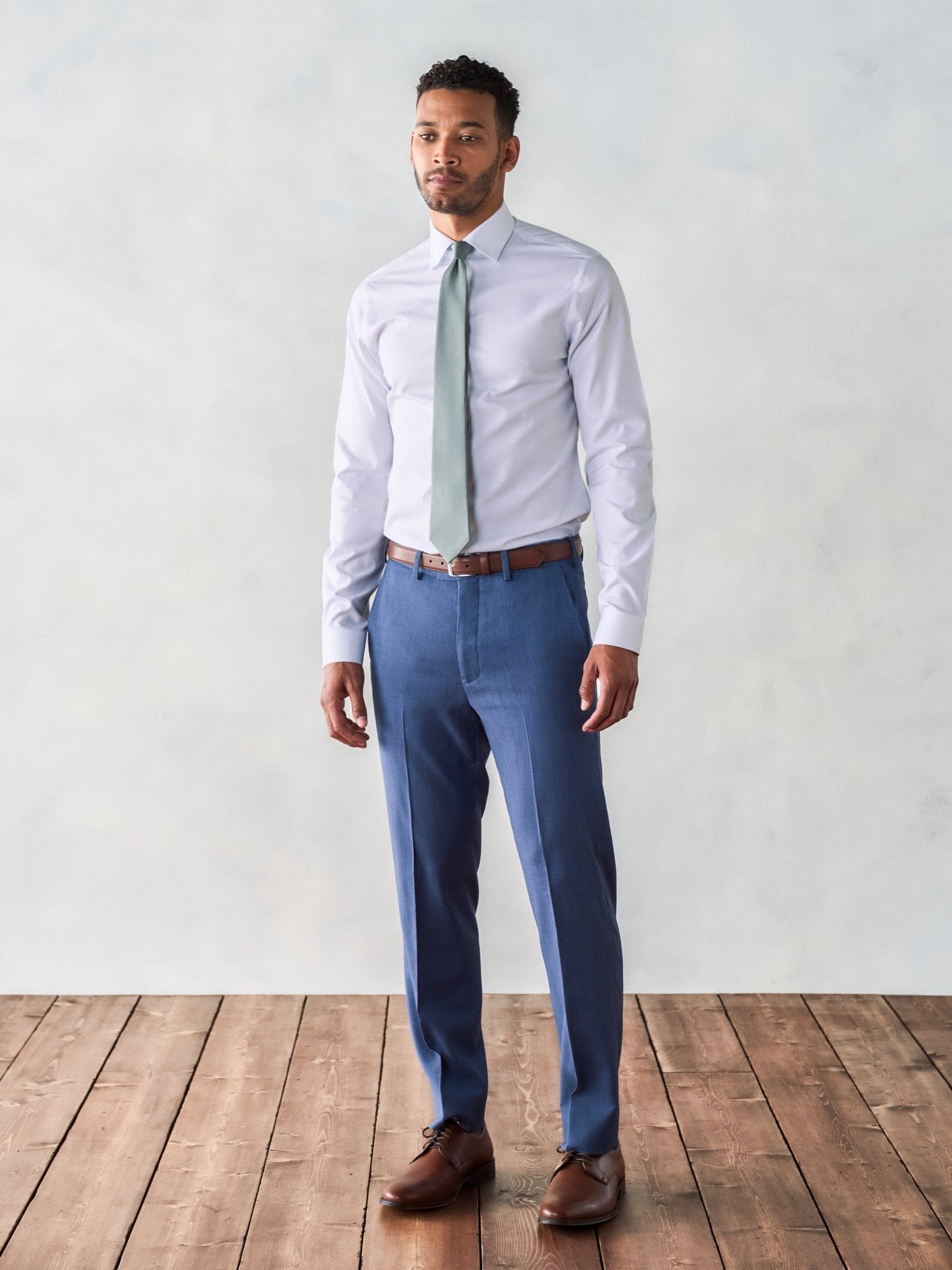 Man in a light shirt and blue pants stands on wooden floor against a light background.