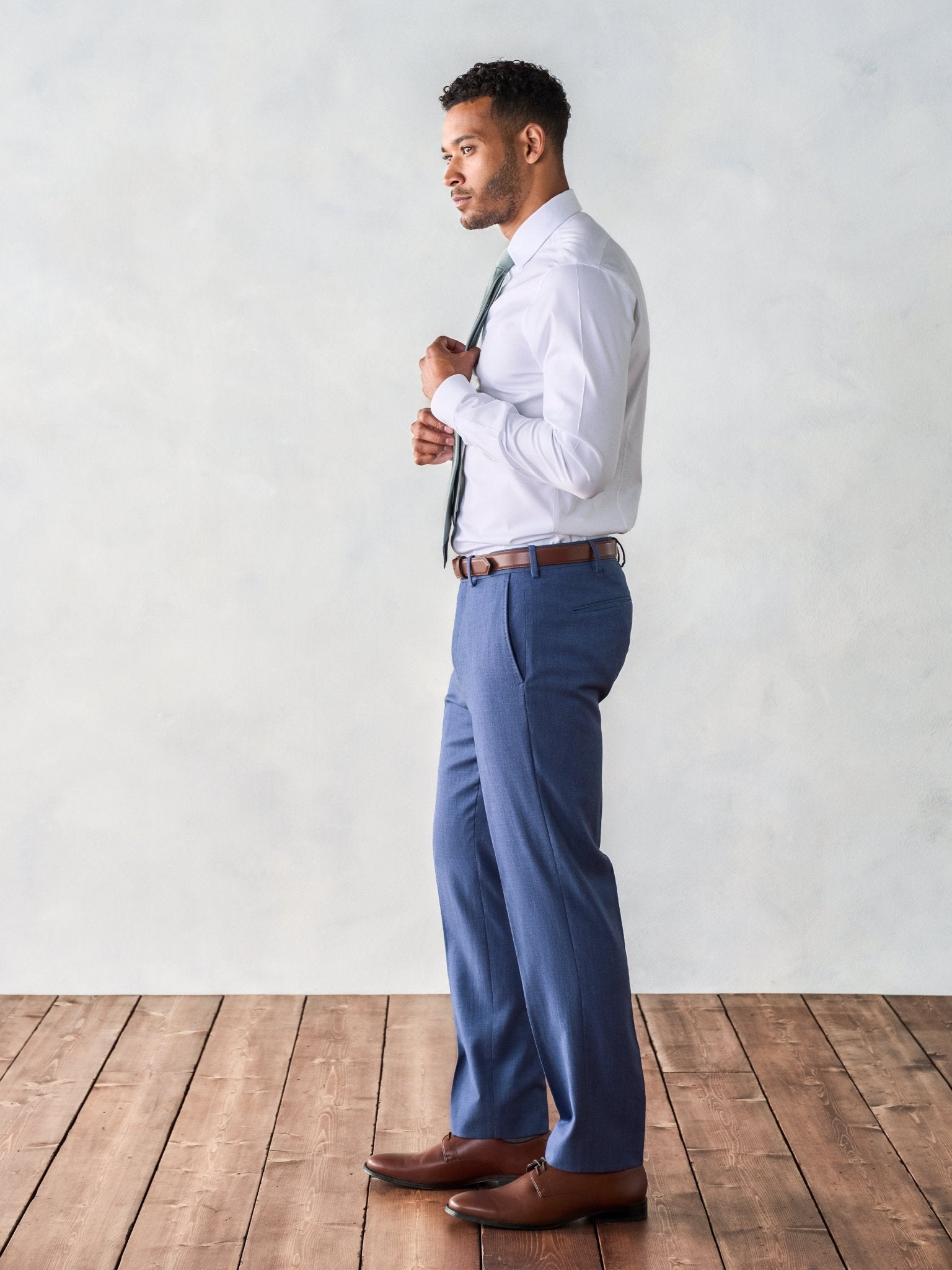 Man in a white shirt and blue pants adjusting his tie, standing on wooden floor.