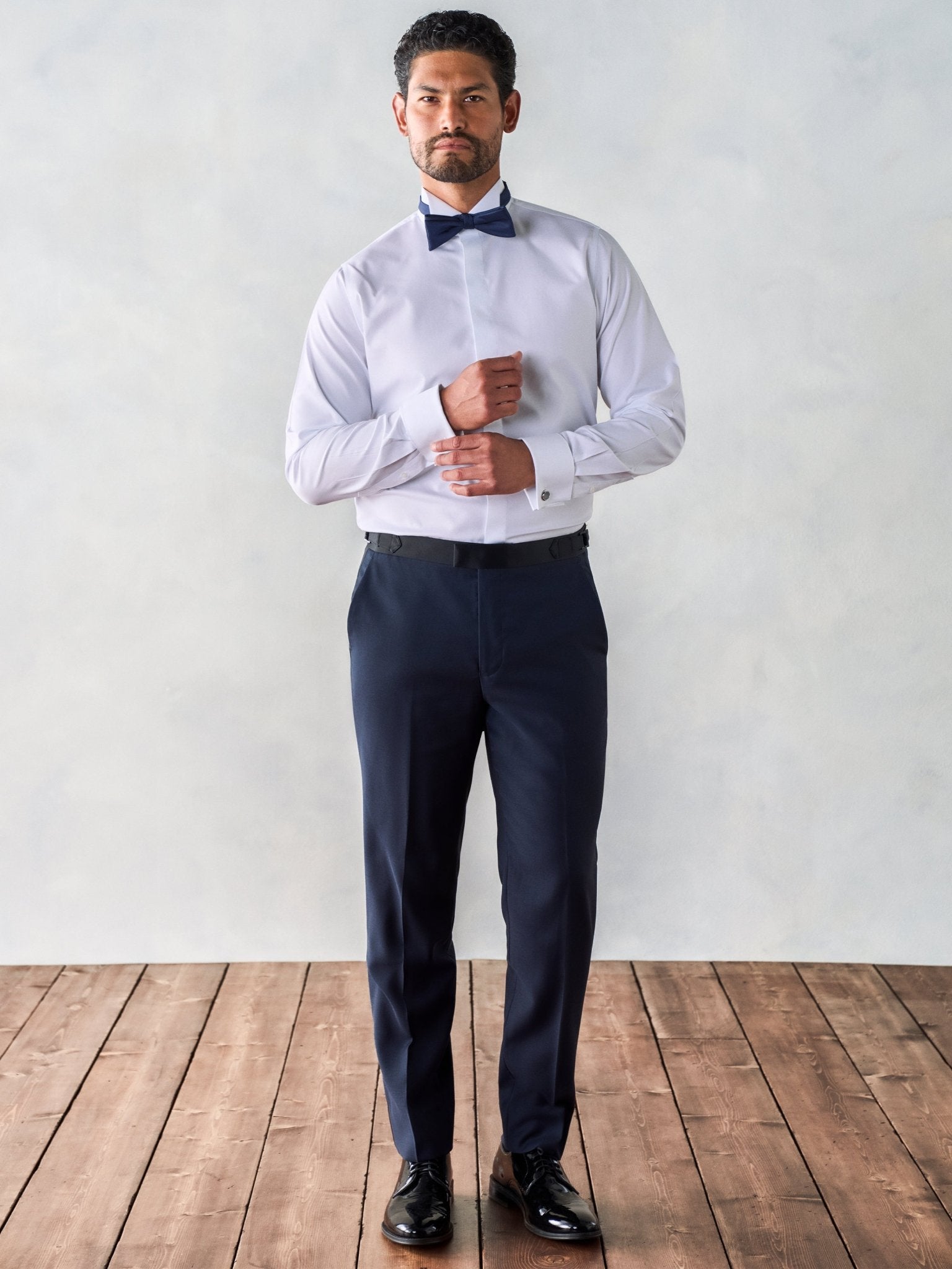 Man in a white dress shirt and navy pants with a bow tie, standing on wooden floor.