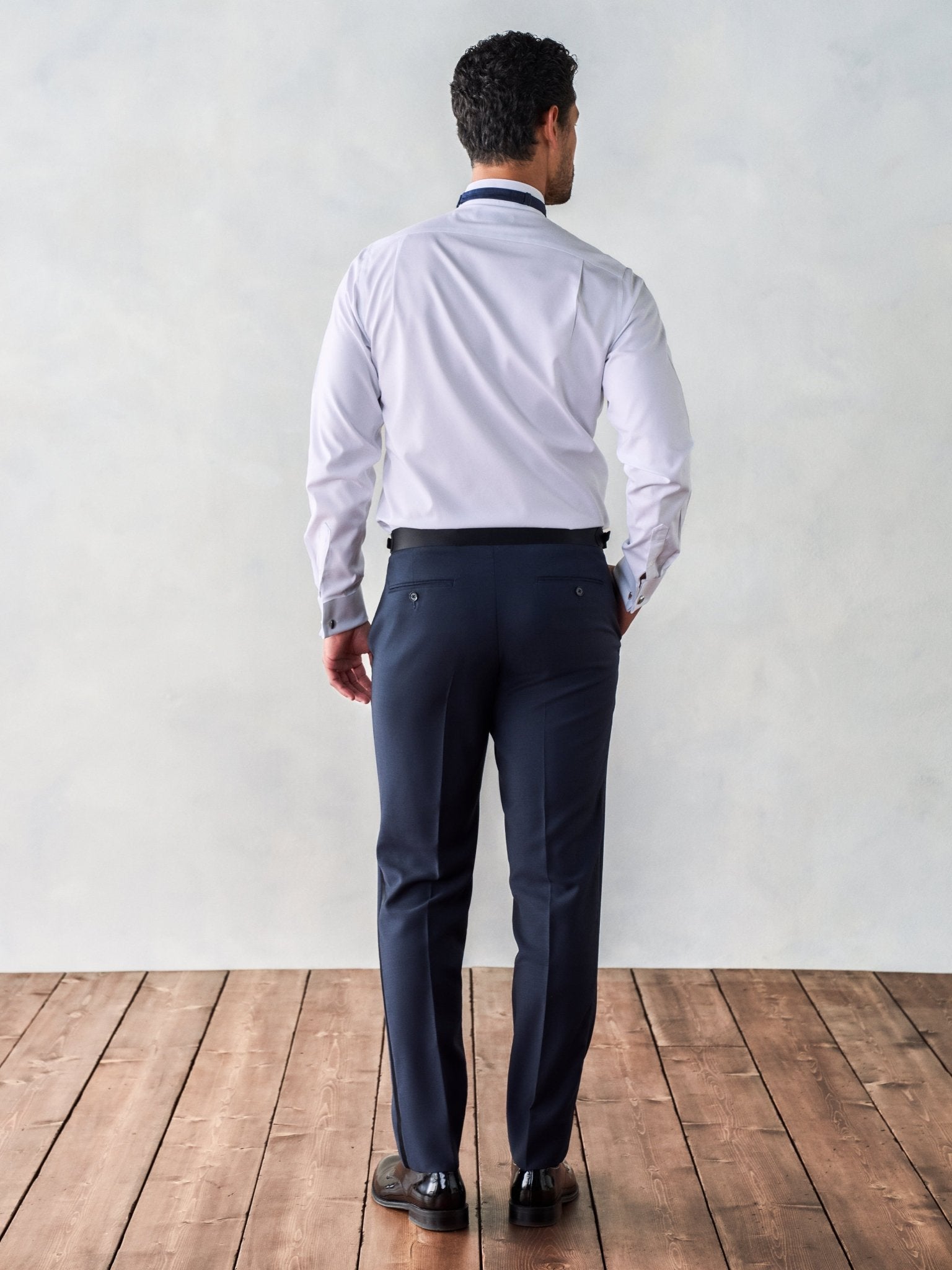 Man in a white shirt and navy pants, viewed from the back, standing on wooden floor.
