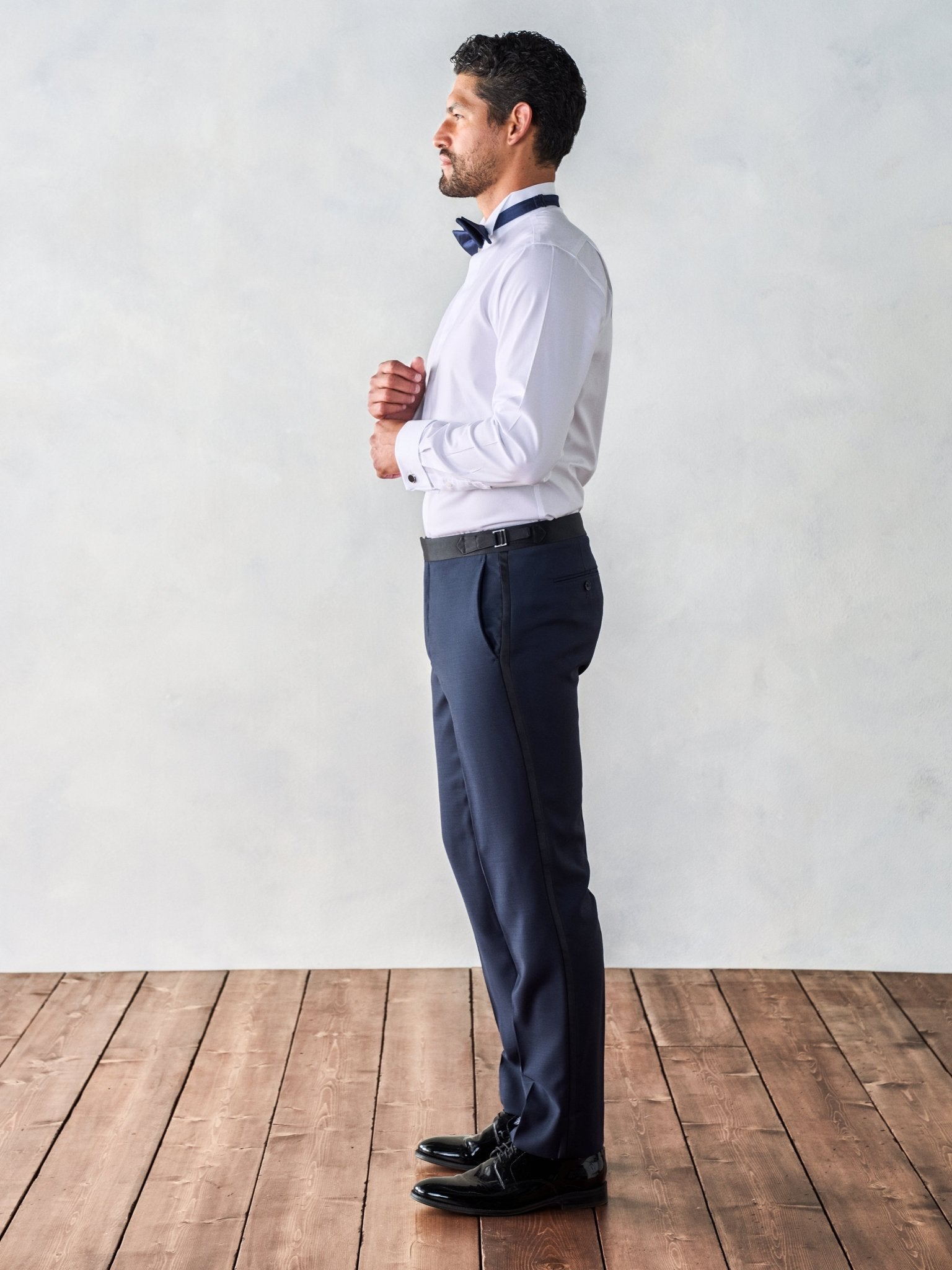 Man in a white shirt and navy pants, adjusting his bow tie, standing on wooden floor.