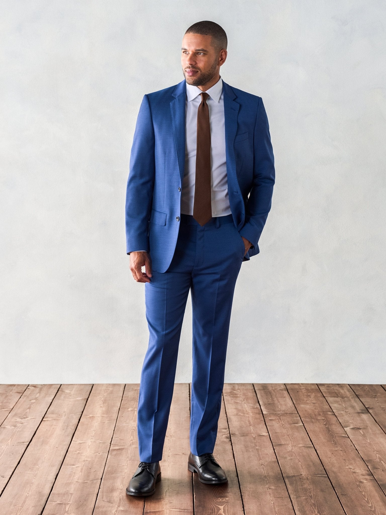 Man in a blue suit with a brown tie, standing on wooden floor against a light background.