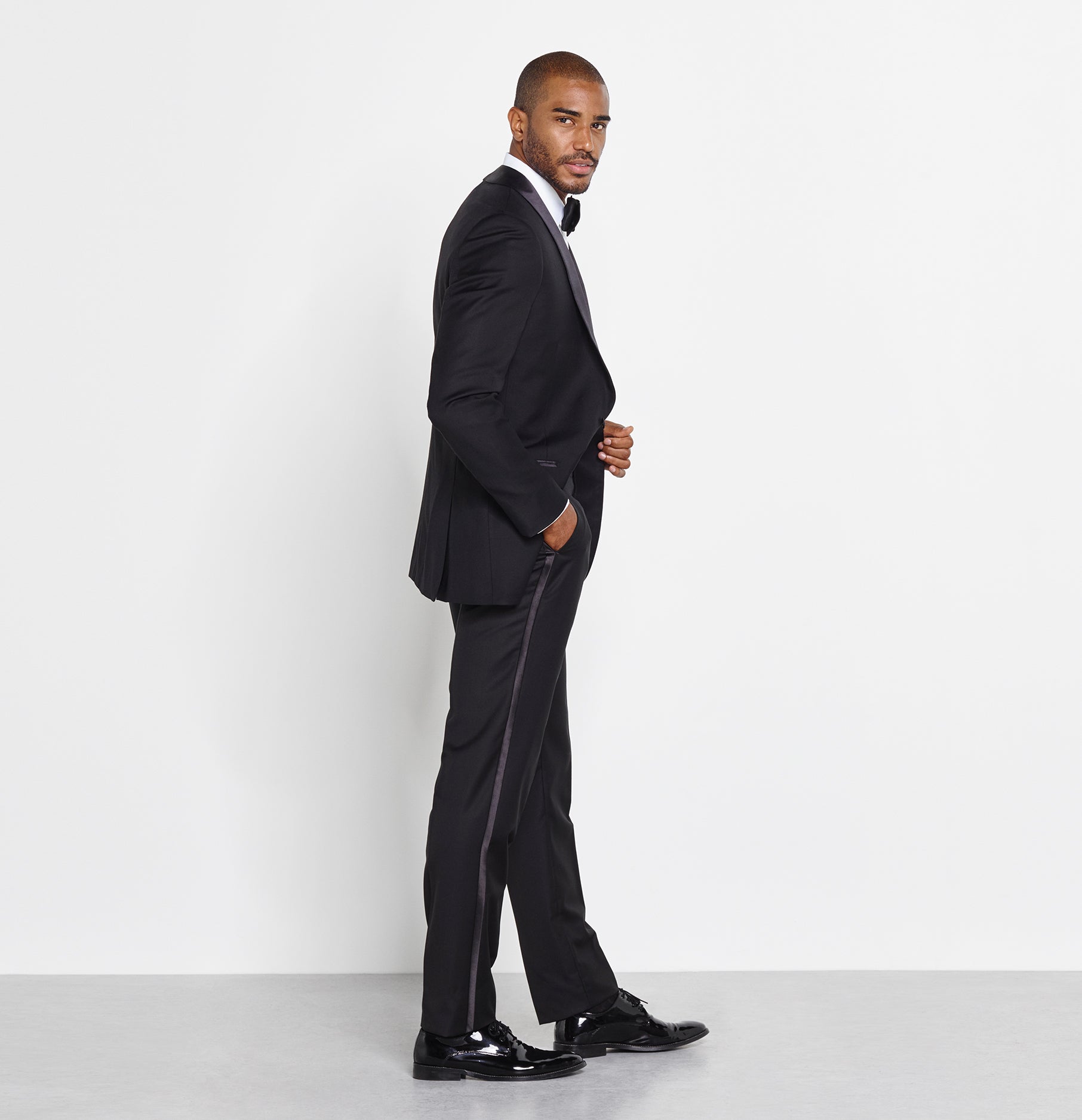 Man in a black tuxedo with a bow tie, standing sideways against a white background.